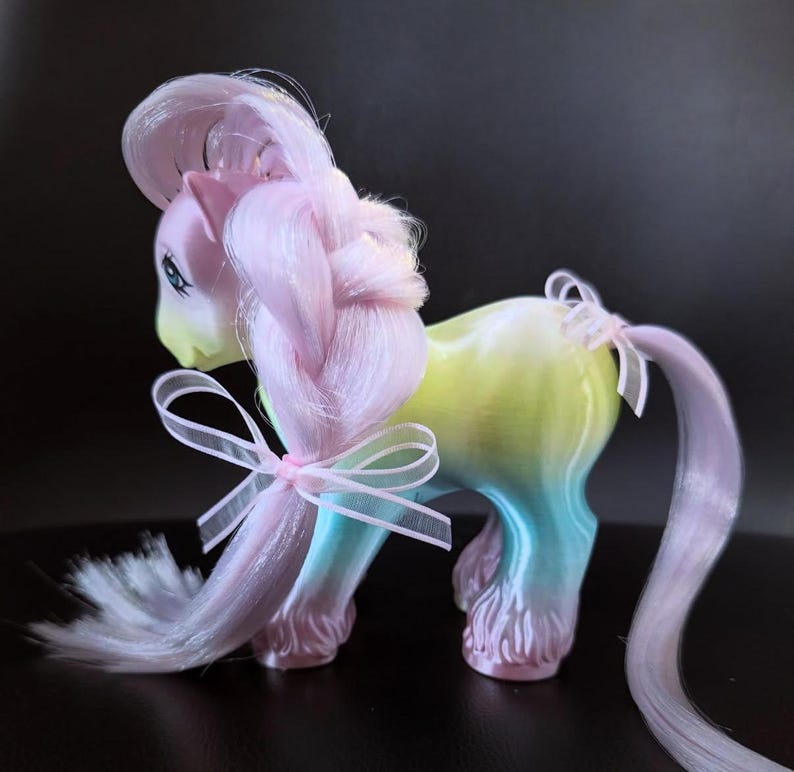 May include: A pastel-colored toy pony with a braided mane and tail, adorned with delicate pink bows. The pony's body transitions from yellow to blue, with pink hooves and a matching pink mane. The toy has blue eyes and a light yellow face.