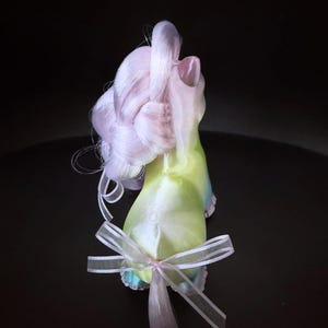 May include: A whimsical, pastel-colored figurine with a flowing, light purple mane and a green and blue body. A delicate white ribbon is tied around the base. The figurine is set against a dark background.