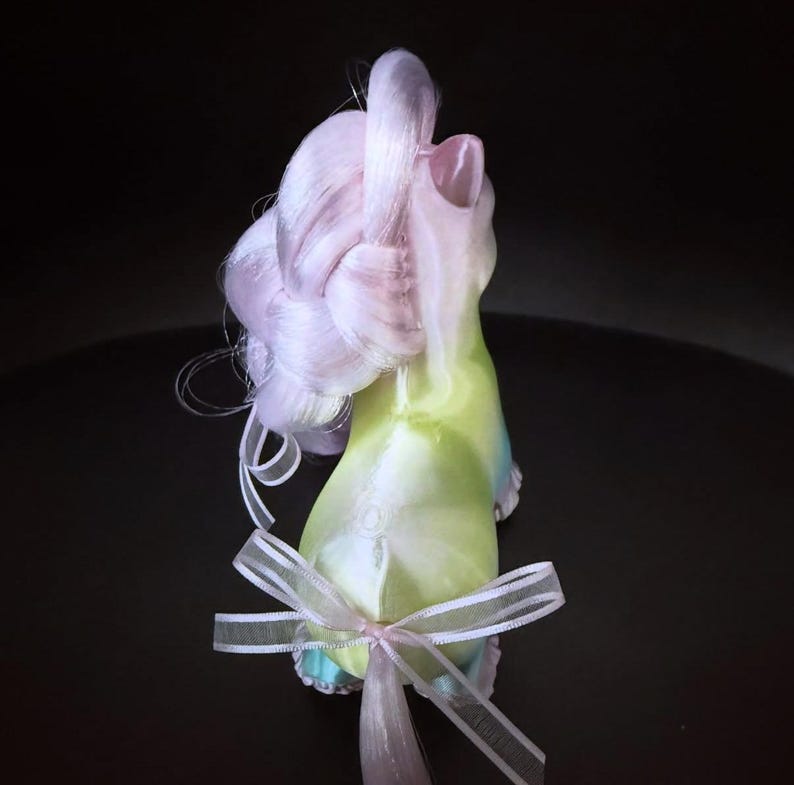 May include: A whimsical, pastel-colored figurine with a flowing, light purple mane and a green and blue body. A delicate white ribbon is tied around the base. The figurine is set against a dark background.