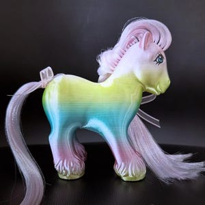 May include: A colorful toy pony figurine with a rainbow body, pink mane, and tail. The pony has blue eyes and is adorned with a white ribbon. The figurine is a collectible item.