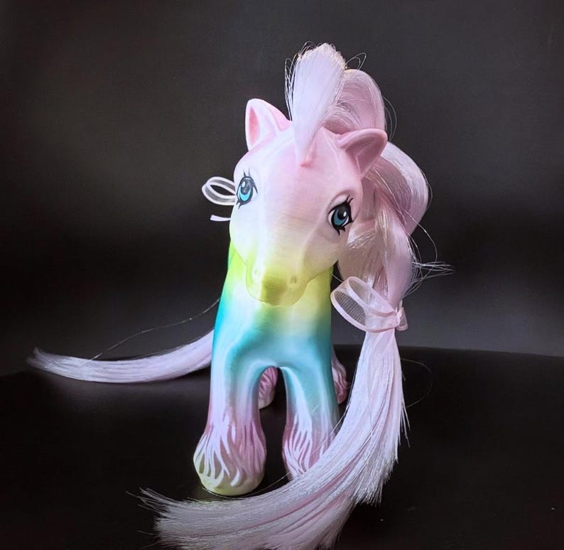 May include: A colorful toy pony with a rainbow gradient body and a pink mane and tail. The pony has blue eyes and a white bow tied around its neck. The toy is a vintage-style collectible.