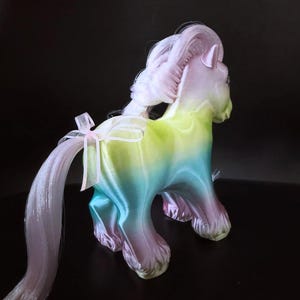 May include: A pastel-colored toy pony with a flowing mane and tail. The pony's body transitions from green to blue to purple. A white ribbon with a pink bow adorns the tail. The toy is set against a dark background.