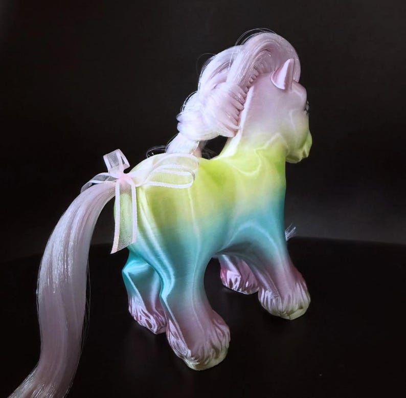 May include: A pastel-colored toy pony with a flowing mane and tail. The pony's body transitions from green to blue to purple. A white ribbon with a pink bow adorns the tail. The toy is set against a dark background.