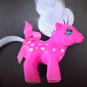 May include: A pink toy pony with a white braided mane and tail. The pony has a pink ribbon tied to its tail and white and yellow hearts on its body. The pony has blue eyes.