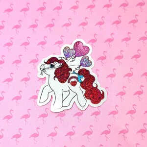 May include: A white cartoon pony sticker with red glitter hair and a red glitter tail. The pony is holding three heart-shaped balloons with glitter accents. A blue ribbon is tied around the pony's neck. The background is pink with flamingo patterns.