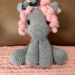 May include: A handmade gray crocheted pony with pink curly hair and a black bow. The pony has black button eyes and is sitting on a pink, textured blanket. The pony's body is gray with silver thread.