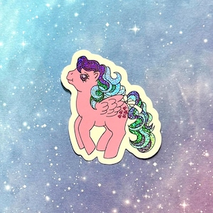 May include: A pink sticker featuring a cartoon pony with a purple, blue, and green mane and tail. The pony has glitter accents and heart-shaped details on its wings. The sticker is on a cream-colored background with a blue and purple starry sky.