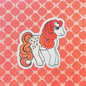 May include: A white pony with a red and orange mane and tail. The pony has a strawberry design on its side.