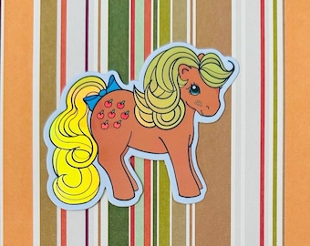 May include: A vintage sticker of a brown pony with a yellow mane and tail. The pony has a blue bow and red apples on its side.
