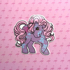 May include: A sticker featuring a purple pony with pink hair and floral accents. The pony has a heart-shaped marking and blue floral designs on its body. The background is a pink pattern of flamingo silhouettes.