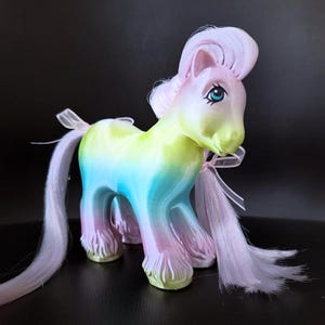 May include: A colorful toy pony with a rainbow gradient body, featuring shades of yellow, blue, and pink. The pony has a pink mane and tail, blue eyes, and a delicate pink ribbon tied around its neck. The toy is set against a dark background.