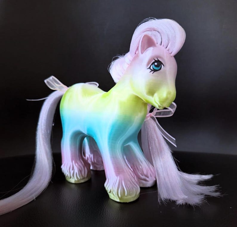 May include: A colorful toy pony with a rainbow gradient body, featuring shades of yellow, blue, and pink. The pony has a pink mane and tail, blue eyes, and a delicate pink ribbon tied around its neck. The toy is set against a dark background.