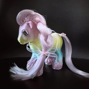 May include: A vintage toy pony with a pastel color scheme. The pony has a pink mane and tail, with a gradient body of yellow, blue, and pink. A pink ribbon is tied around the tail and neck. The pony has blue eyes and is posed on a dark surface.
