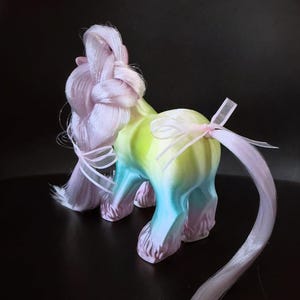 May include: A whimsical, colorful unicorn figurine with a gradient body of yellow, green, and blue. The unicorn has a pink braided mane and tail, adorned with delicate ribbons. The figurine is set against a dark background.