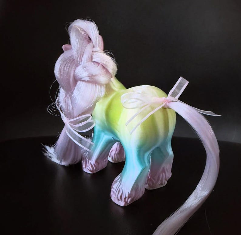 May include: A whimsical, colorful unicorn figurine with a gradient body of yellow, green, and blue. The unicorn has a pink braided mane and tail, adorned with delicate ribbons. The figurine is set against a dark background.