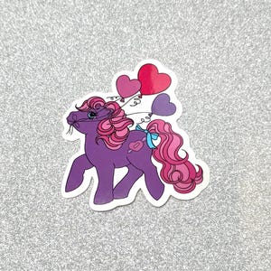 May include: A purple pony sticker with pink hair and a blue bow. The pony has a heart-shaped design on its side and is holding three heart-shaped balloons. The sticker is on a silver glitter background.
