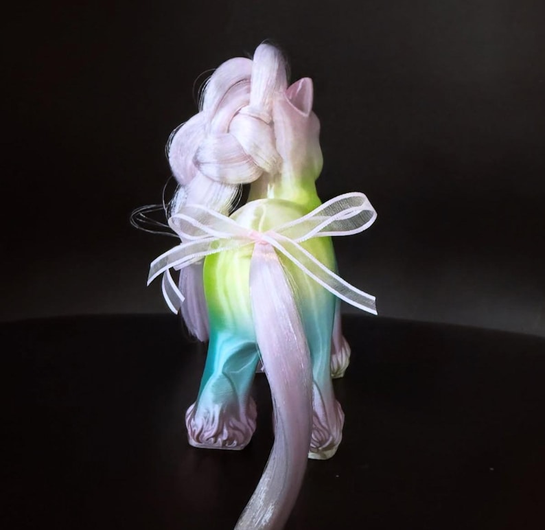 May include: A colorful unicorn figurine with a gradient body of green, blue, and purple. The unicorn has a long, braided mane and tail in shades of pink and white, tied with a sheer white ribbon. The figurine is on a dark surface.