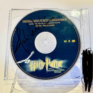 Może przedstawiać: Podpisana płyta CD ze ścieżką dźwiękową do filmu Harry Potter i więzień Azkabanu. Płyta CD znajduje się w przezroczystym plastikowym etui. Płyta CD ma niebieskie tło z białym sową latającą w lewym górnym rogu. Tekst "Harry Potter i więzień Azkabanu" jest w żółtych i białych literach. Tekst "Oryginalna ścieżka dźwiękowa filmu" jest w białych literach. Tekst "Muzyka skomponowana i dyrygowana przez Johna Williamsa" jest w białych literach.
