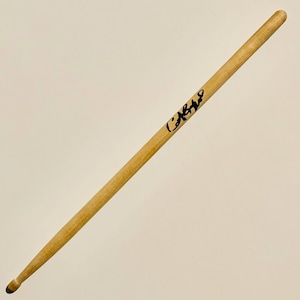 May include: A single wooden drumstick with a rounded tip. The drumstick is light brown and has a black signature on it. The drumstick is angled from the top left to the bottom right.