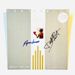 Pode incluir: Uma capa quadrada de disco de vinil para Eurythmics Sweet Dreams are made of this. A capa apresenta uma imagem central de uma pessoa segurando um coração vermelho, com duas assinaturas distintas em tinta azul e preta sobre um fundo cinza com barras douradas.