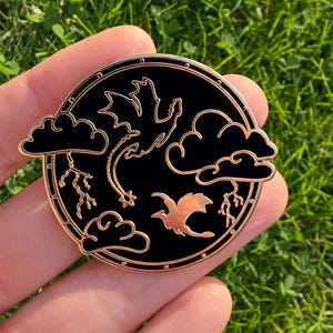 May include: Round black and gold enamel pin featuring a dragon design with clouds and lightning. The pin has a gold border and a detailed illustration of dragons in flight. A unique accessory for fantasy enthusiasts.