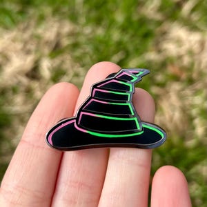 May include: A black witch hat enamel pin with pink and green outlines. The hat has a pointed top and a wide brim.