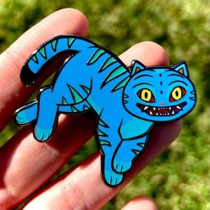 May include: A blue cat enamel pin with a cartoon design. The cat has yellow eyes, a wide grin, and green stripes. The pin is held in a hand against a blurred green background.