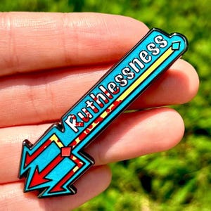 Peut inclure: Épingle en émail turquoise et rouge avec le mot "Ruthlessness" en lettres blanches. L'épingle a une bande jaune et des détails de flèches rouges. L'épingle est tenue dans une main sur un fond vert flou.