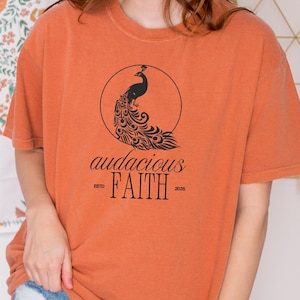 May include: Orange t-shirt with a black peacock design inside a circle and the words "audacious FAITH" below. The shirt is a solid orange color and has short sleeves. The model is wearing blue jeans.