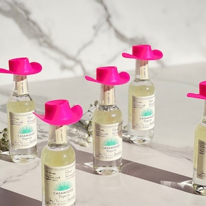 May include: Five miniature bottles of Casamigos Tequila, each topped with a pink cowboy hat. The bottles are clear glass and the labels are white with green text.