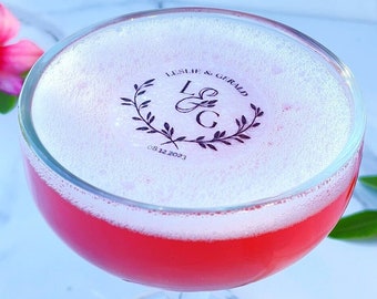 May include: A pink cocktail in a stemmed glass with a white foam top. The foam has a black design with the text "Leslie & Gerald L&G 08.12.2023".