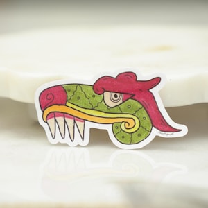 May include: A die-cut sticker featuring a stylized illustration of a serpent head with a green textured body, a red crest, a yellow jawline, and sharp white teeth. The design includes a single visible eye and a curled tail section.