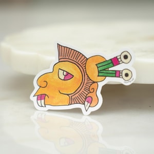 May include: A die-cut sticker featuring a stylized profile illustration of a figure with yellow skin, a brown headdress, and two green and pink cylindrical ornaments extending from the back of the head. The design includes black outlines.