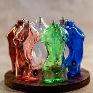 May include: Five glass figurines in the shape of a human torso, each a different color: pink, green, clear, blue, and light blue. The figurines are arranged in a circle on a wooden base.