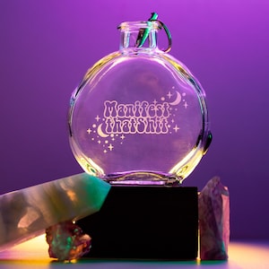 May include: A clear glass bottle with a round shape and a silver lid. The bottle has the words "Manifest that shit" etched into the glass, along with stars and crescent moons. The bottle is sitting on a black surface with a white and green stone in the foreground.