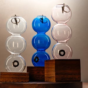 May include: Three glass sphere ornaments stacked on wooden bases. The top sphere is pink, the middle sphere is blue, and the bottom sphere is clear. Each sphere has a small hole in the bottom.