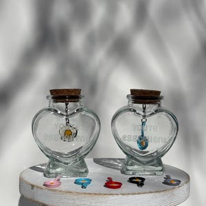 May include: Two clear, heart-shaped glass jars with cork stoppers. Each jar has a charm inside and the words "You're eggcellent" printed on the front. Several small, colorful egg-shaped charms are on a white wooden surface.