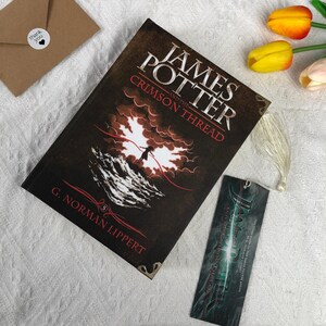 May include: A hardcover book titled "James Potter and the Crimson Thread" with a matching bookmark. The book cover is dark with a dramatic illustration of a figure against a red and white background. A small "thank you" sticker is visible.