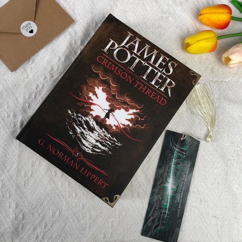 May include: A hardcover book titled "James Potter and the Crimson Thread" with a matching bookmark. The book cover is dark with a dramatic illustration of a figure against a red and white background. A small "thank you" sticker is visible.