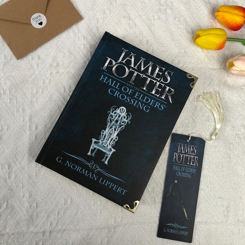 May include: A hardcover book and matching bookmark with the title "James Potter and the Hall of Elders' Crossing." The book has a dark blue cover with silver lettering and a detailed illustration of a chair. A small "thank you" card is also present.