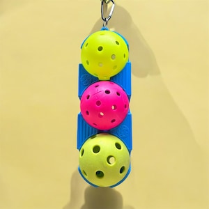 Könnte beinhalten: Ein blaues und gelbes Hängespielzeug mit drei bunten Bällen. Die Bälle sind gelb, rosa und grün und haben Löcher. Das Spielzeug ist für Vögel gedacht.