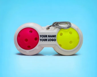 May include: A white pickleball holder with a silver carabiner clip. The holder contains a pink and a yellow pickleball. The text "YOUR NAME YOUR LOGO" is printed on the holder. The background is a gradient of blue.