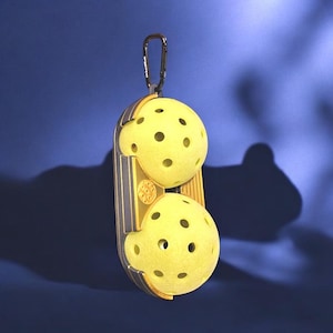 Puede incluir: Un soporte de pickleball dorado y gris que contiene dos pelotas de pickleball amarillas. Las pelotas tienen múltiples agujeros. El soporte tiene un mosquetón. El fondo es un degradado de azul.