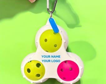 May include: A white pickleball holder with three balls: two yellow and one pink. The holder has a carabiner clip and a blue tag. Text on the holder reads "YOUR NAME YOUR LOGO". The background is a solid green.