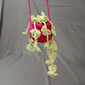 May include: A handmade, crochet hanging planter in bright pink, filled with light green crochet foliage. The planter is suspended by a matching pink cord. The leaves cascade down, creating a whimsical, decorative piece for indoor plants.