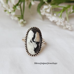 Pode incluir: Anel oval com uma pedra preta e branca em uma banda de prata. A pedra tem um padrão único. O anel está em uma superfície branca com detalhes florais. O texto "Shreejaipurjewels92" está na parte inferior.