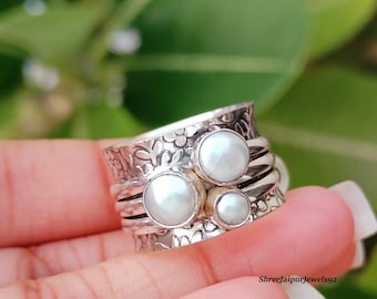 Könnte beinhalten: Ein silberner Spinner-Ring mit drei weißen Perlen. Der Ring hat ein Blumenmuster auf dem Band und wird in einer Hand vor einem verschwommenen grünen Hintergrund gehalten. Das Design des Rings umfasst mehrere rotierende Bänder.