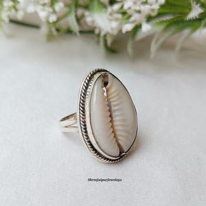 Puede incluir: Anillo de plata con una concha de cauri grande y ovalada. La concha es de color blanco crema con marcas marrones, engastada en una banda de plata con un diseño de cuerda trenzada. El texto "Shreejaipurjewels92" es visible.