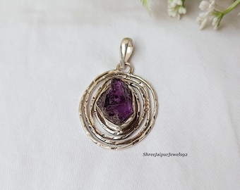 May include: A silver pendant featuring a raw, deep purple amethyst gemstone. The stone is set within a textured, circular frame with concentric rings. The pendant has a bail for a chain. The background is a white fabric.