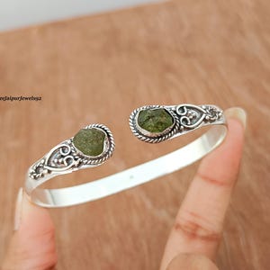May include: A silver cuff bracelet featuring two raw, olive-green stones set at each open end. The metal band displays intricate, swirling wirework patterns surrounding the stones. The text ShreeJaipurJewels92 is visible on the left side.